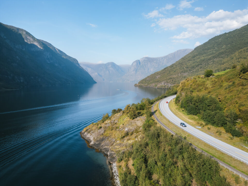 Mietwagen fährt auf einer Panoramastraße an einem norwegischen Fjord mit einem Boot im Hintergrund